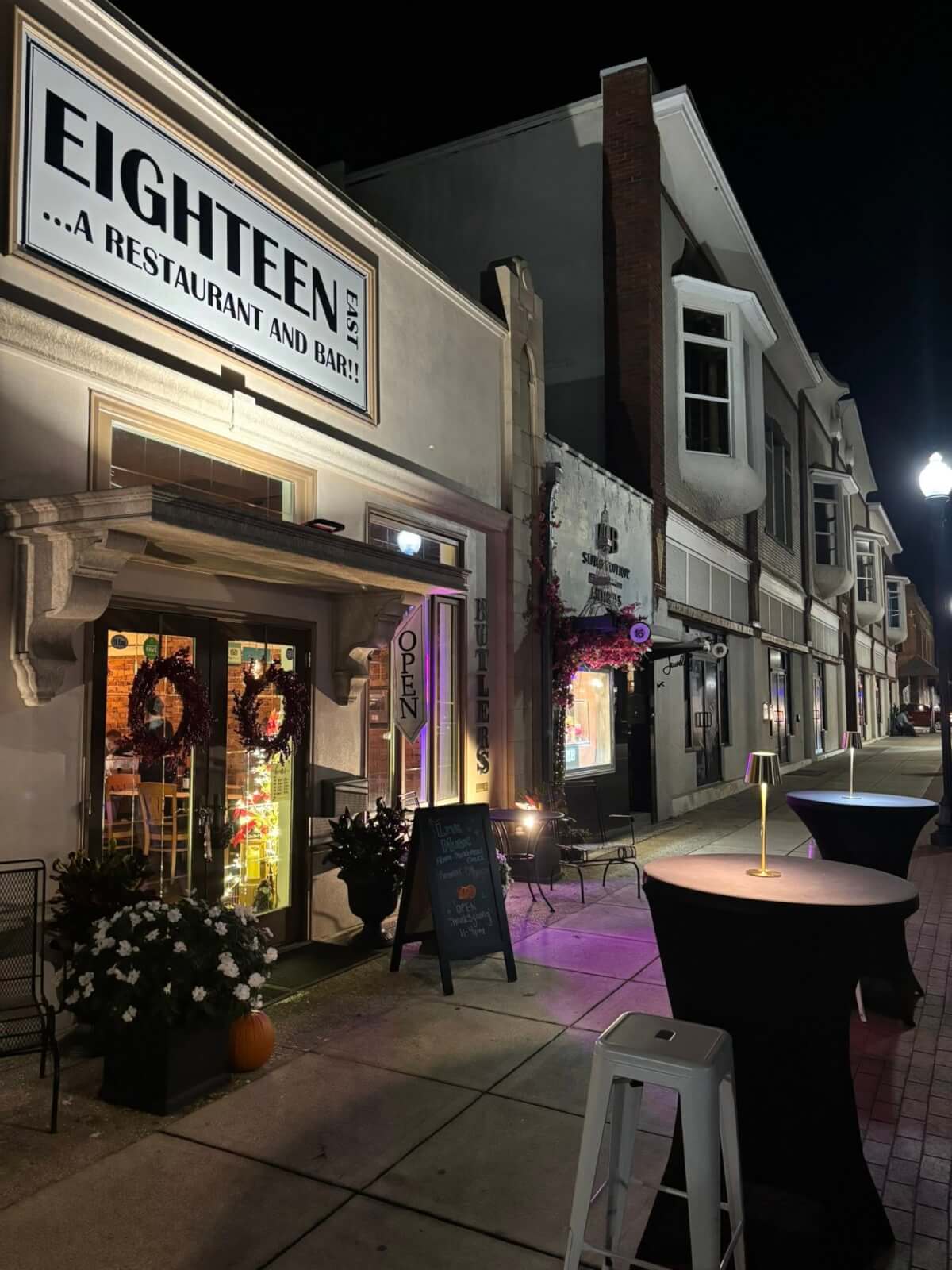 Nighttime exterior of 18 East Restaurant and Bar in Avon Park, Florida, with a lit storefront sign, glass entrance doors decorated with wreaths, potted flowers by the doorway, and tall cocktail tables with small lamps set up along a quiet downtown sidewalk.