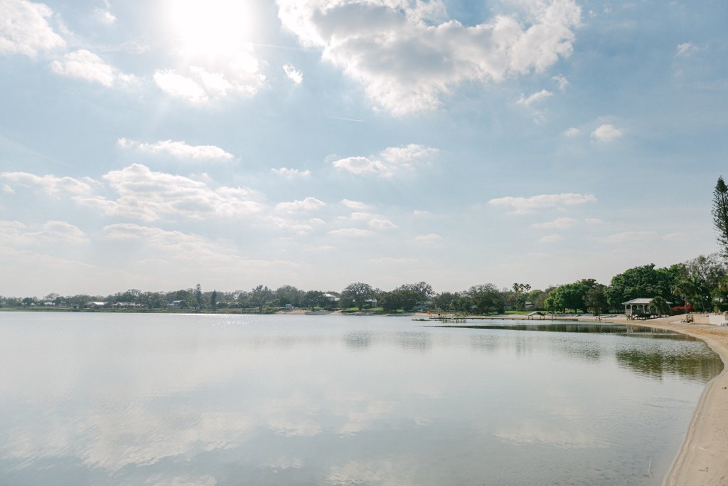 2023 02 24 LakeTulane AvonParkFL 0005-visit-sebring-fl A still lake is reflecting a light blue sky with puffy white clouds. Green trees, docks and houses dot the shoreline around the lake.