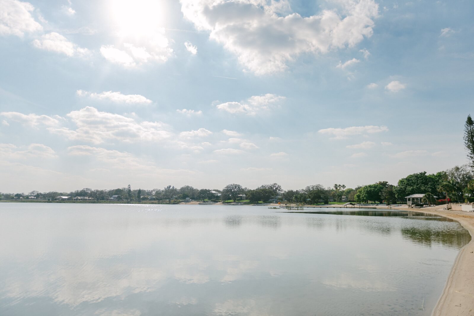 A still lake is reflecting a light blue sky with puffy white clouds. Green trees, docks and houses dot the shoreline around the lake.