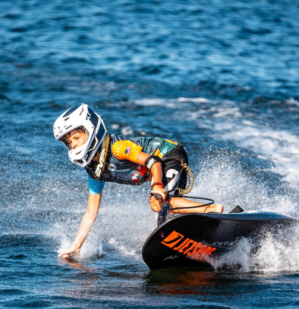 A young boy wearing a helmet and safety gear leans into a sharp turn while motosurf racing on Lake Jackson in Sebring, Florida, with water spraying around him.