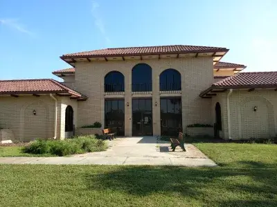 AP community center-visit-sebring-fl Exterior view of a tan brick building with a red tile roof and three large arched windows centered above the entrance. The building has symmetrical wings on each side, two benches near the doorway, and a walkway leading across a lawn under a clear blue sky.