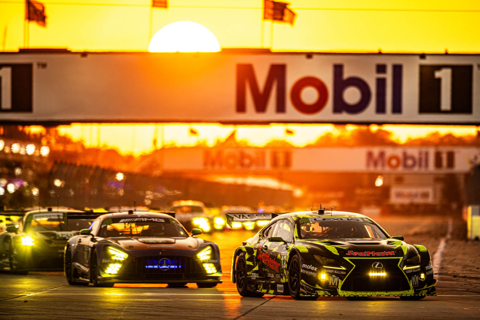 Lexus and Mercedes-AMG race cars compete beneath the Mobil 1 bridge at sunset at Sebring International Raceway.