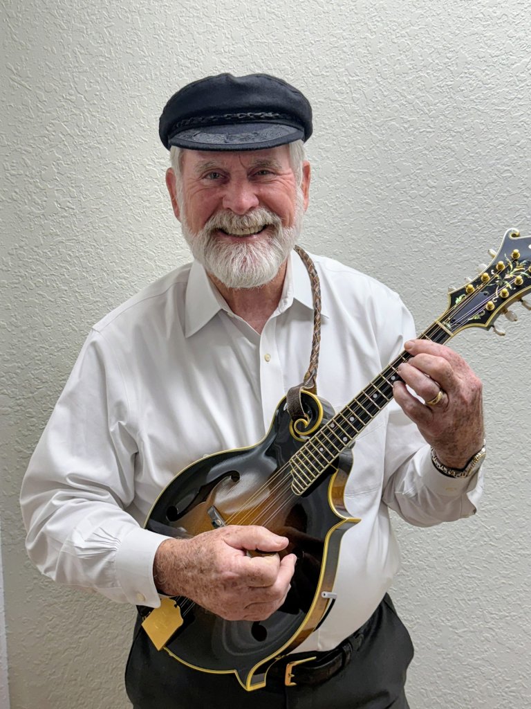 Smiling older man with a white beard, wearing a white shirt and black cap, holding and playing a mandolin against a light-colored wall.
