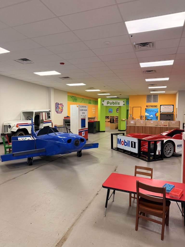 Interior play area showcasing a blue airplane and Mobil 1 race car that can be sat inside of with a USPS carrier truck in the back ground.