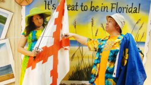 A man and woman sword fighting while wearing strange hats in front of a Florida poster