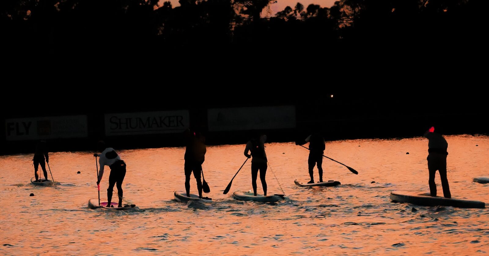 Silhouettes of several stand-up paddleboarders glide through glowing sunset water, capturing the endurance and determination that define the Last Paddler Standing competition, taking place December 6–8 in Lake Placid, Florida