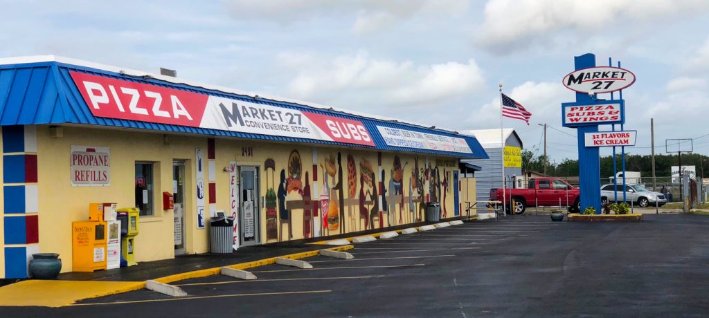 Market 27 in Avon Park, FL with blue awning, wall mural of diners, and sign for pizza, subs, wings & ice cream.