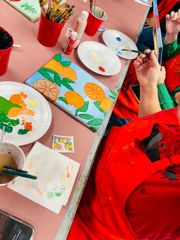 Photo shows an artist painting an image of orange slices and whole fruits with green leaves atop a light blue canvas as part of a painting class hosted by The Red Easel in Sebring, FL