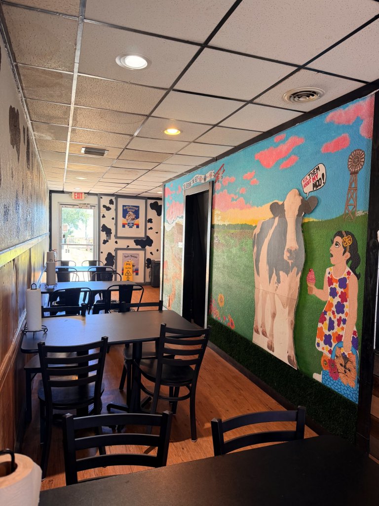 Inside seating area with black tables and chairs and to the right a mural of a little girl holding an ice cream looking up at a cow.