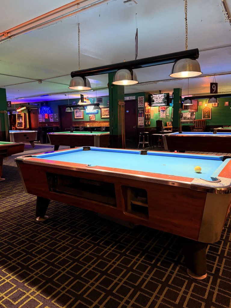 bc-visit-sebring-fl Vibrant pool hall in downtown Sebring featuring green accent walls, neon lighting, a TouchTunes jukebox, bar décor, and five of twelve clean, well-maintained pool tables.