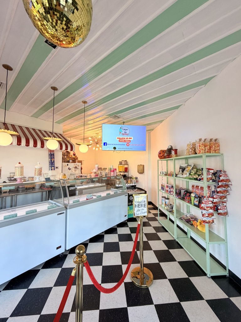 Ice cream shop interior featuring warm lighting, a fully stocked snack shelf, classic black-and-white checkered flooring, and a disco ball hanging from the ceiling for a fun, retro atmosphere.