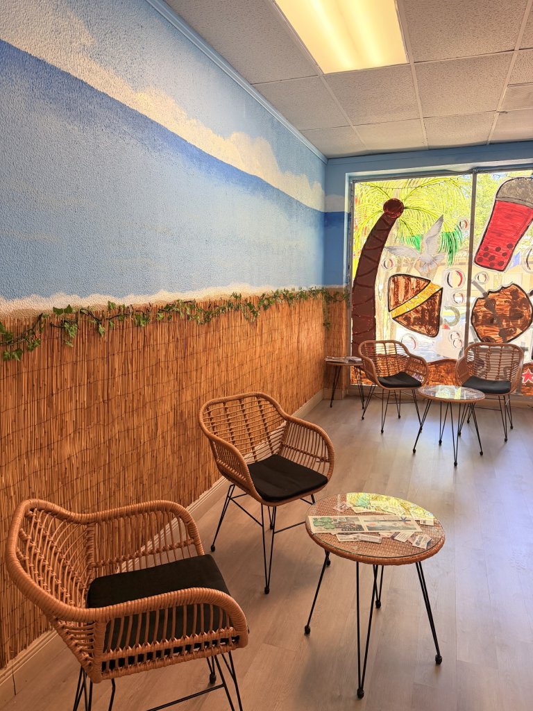 Interior sitting area of boba shop showing 4 rattan chairs with cafe tables against a beach like wall.