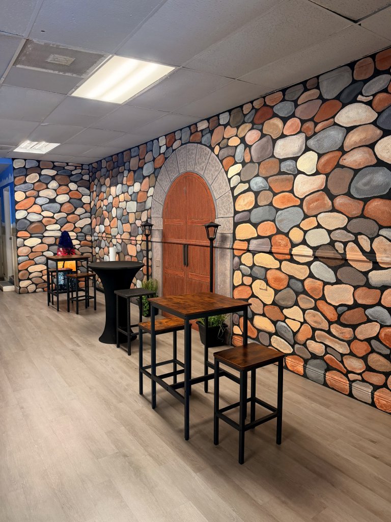 A cozy indoor space with a decorative stone-patterned wall mural featuring an arched wooden door design. The room has light wood flooring and is furnished with tall wooden tables and stools, along with a small round cocktail table.
