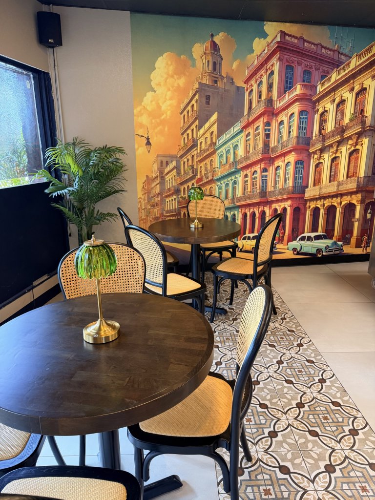 mural back drop of colorful Havana buildings, more cafe style seating, green plants, and wicker chairs