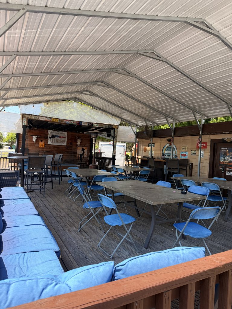 Pictured is some of the exterior of the restaurant showing a large covered patio under a metal roof, blue patio seating along with tables and chairs and an outside stage.