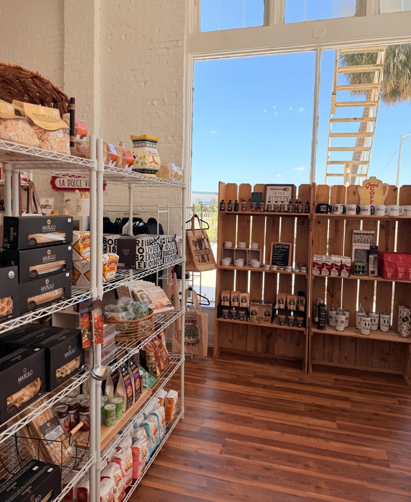 Left metal shelf showcasing variety of pastas, wooden floors line the flooring, and in front is a wooden shelf showing multiple glass products.