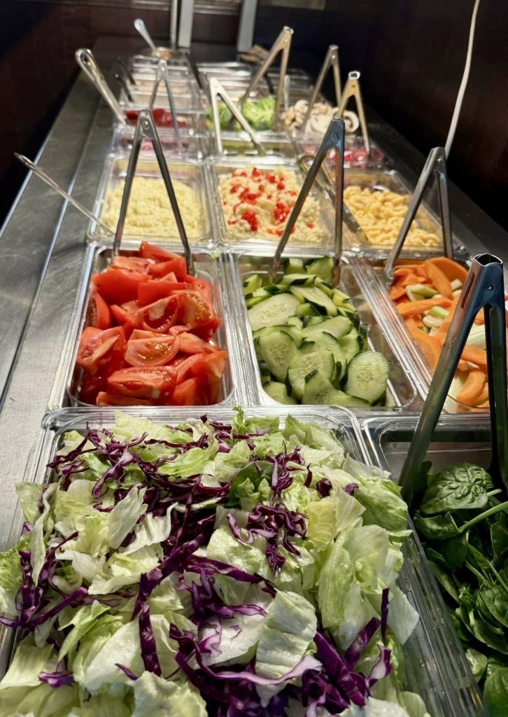 A salad bar showcasing it's wide variety of salad options (pictured) include iceberg lettuce, cut tomatoes, sliced cucumbers, spinach and much more.