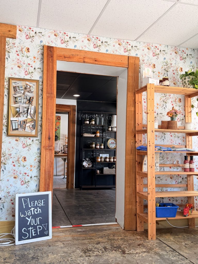 org nat-visit-sebring-fl A cozy, rustic café or boutique interior with floral wallpaper, exposed wood trim, and warm natural textures. The first photo shows a doorway framed in thick reclaimed wood leading into a darker back room with shelving stocked with jars and products. A handwritten chalkboard sign on the floor reads “Please Watch Your Step.” Wooden shelves with plants, folded shirts, baskets, and bottles decorate the right side.