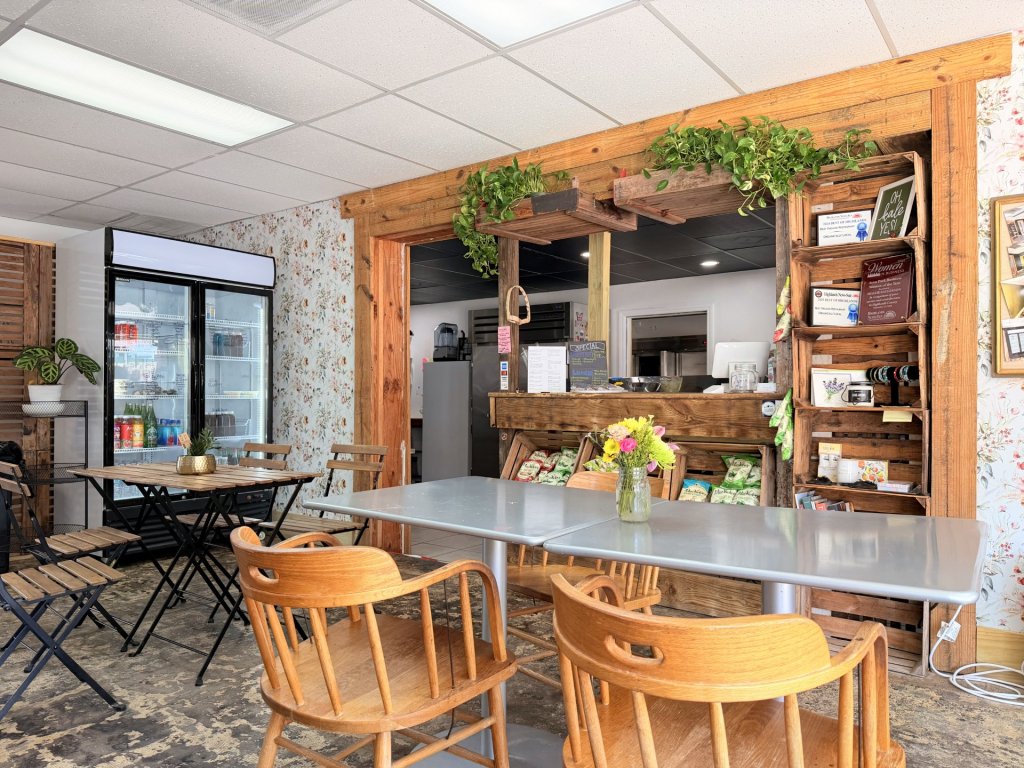 org natt-visit-sebring-fl Wooden and black metal café tables and chairs sit on a worn concrete floor, with flowers in a vase on the center table. A rustic wooden counter framed by reclaimed wood and hanging plants serves as the focal point. Shelves display snacks, products, and awards, while a glass refrigerator stocked with drinks stands to the left. The space has a charming farmhouse-inspired aesthetic with bright, inviting lighting.