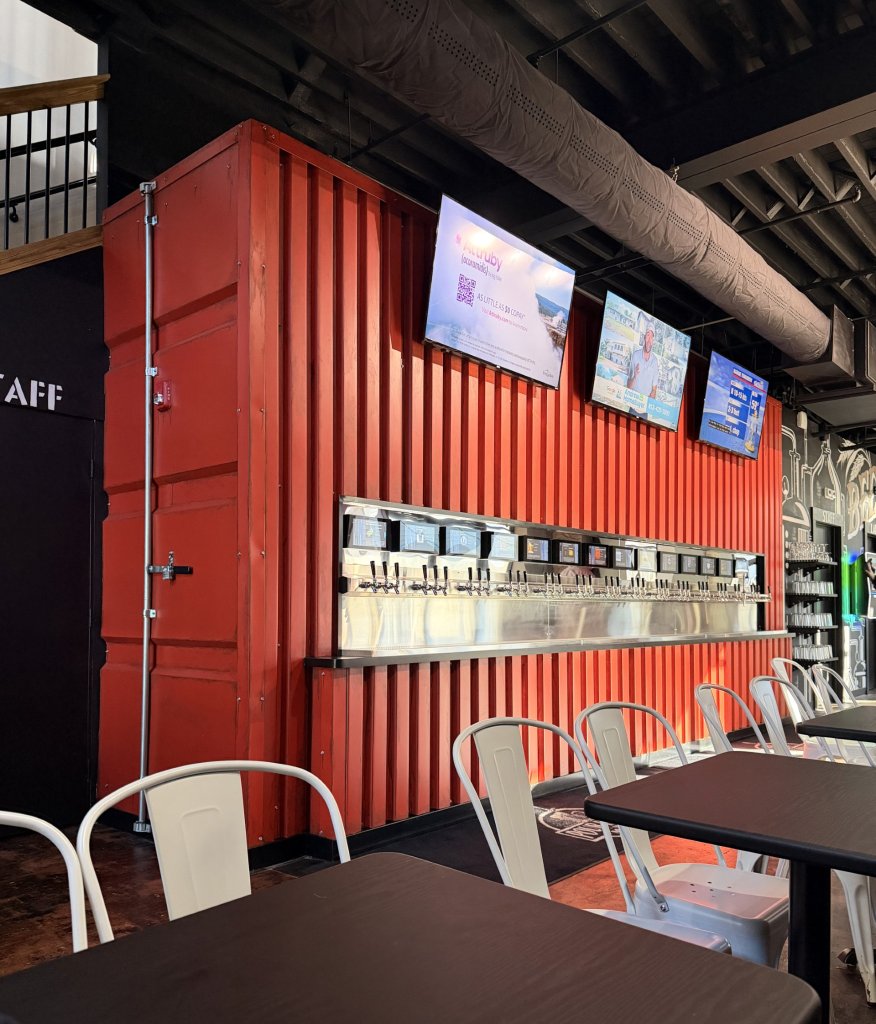 pour housee-visit-sebring-fl Industrial self-pour taproom featuring a red shipping container bar with multiple beer taps, digital screens above, stainless steel draft system, metal chairs, and dark wood tables in a modern brewery interior.