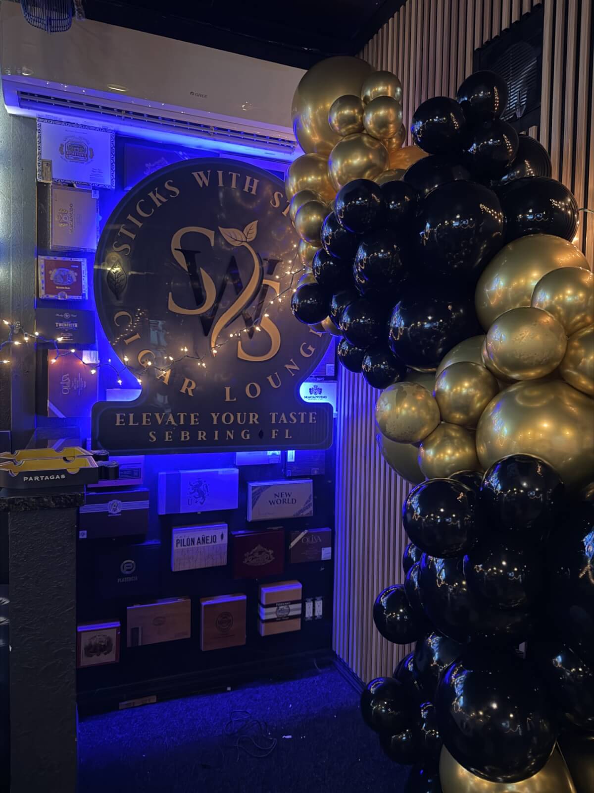 stickss-visit-sebring-fl A warm, upscale cigar lounge corner glowing with blue light, decorated with premium cigar boxes and a bold black-and-gold balloon arch in downtown Sebring.