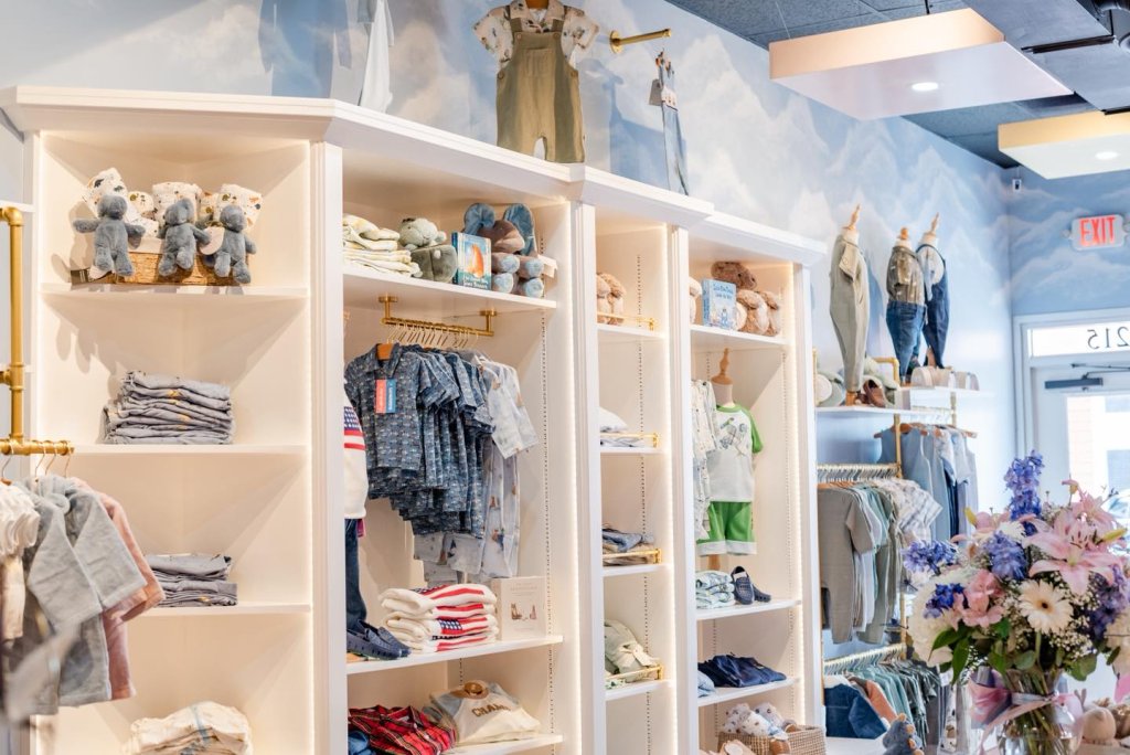 Interior display of a Sebring, FL baby boutique with a blue accent wall decorated with floral elements, showcasing baby boy plush toys, clothing sets, and tiny shoes.