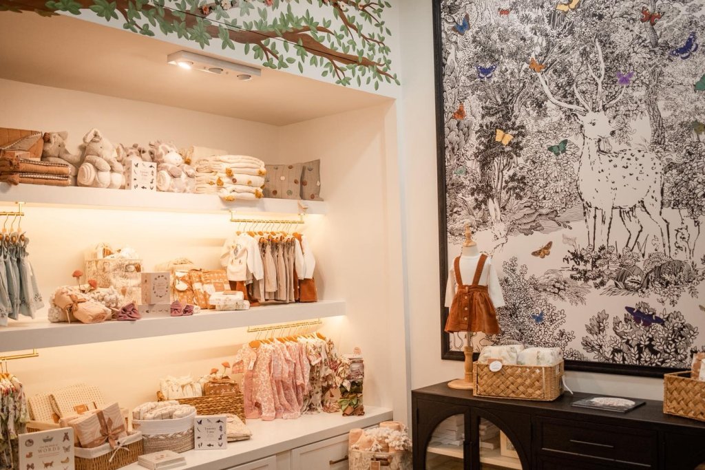 “Interior of a baby boutique in Sebring, FL featuring neatly arranged baby clothes, plush toys, blankets, and accessories on illuminated shelves beside a large woodland mural with a deer.