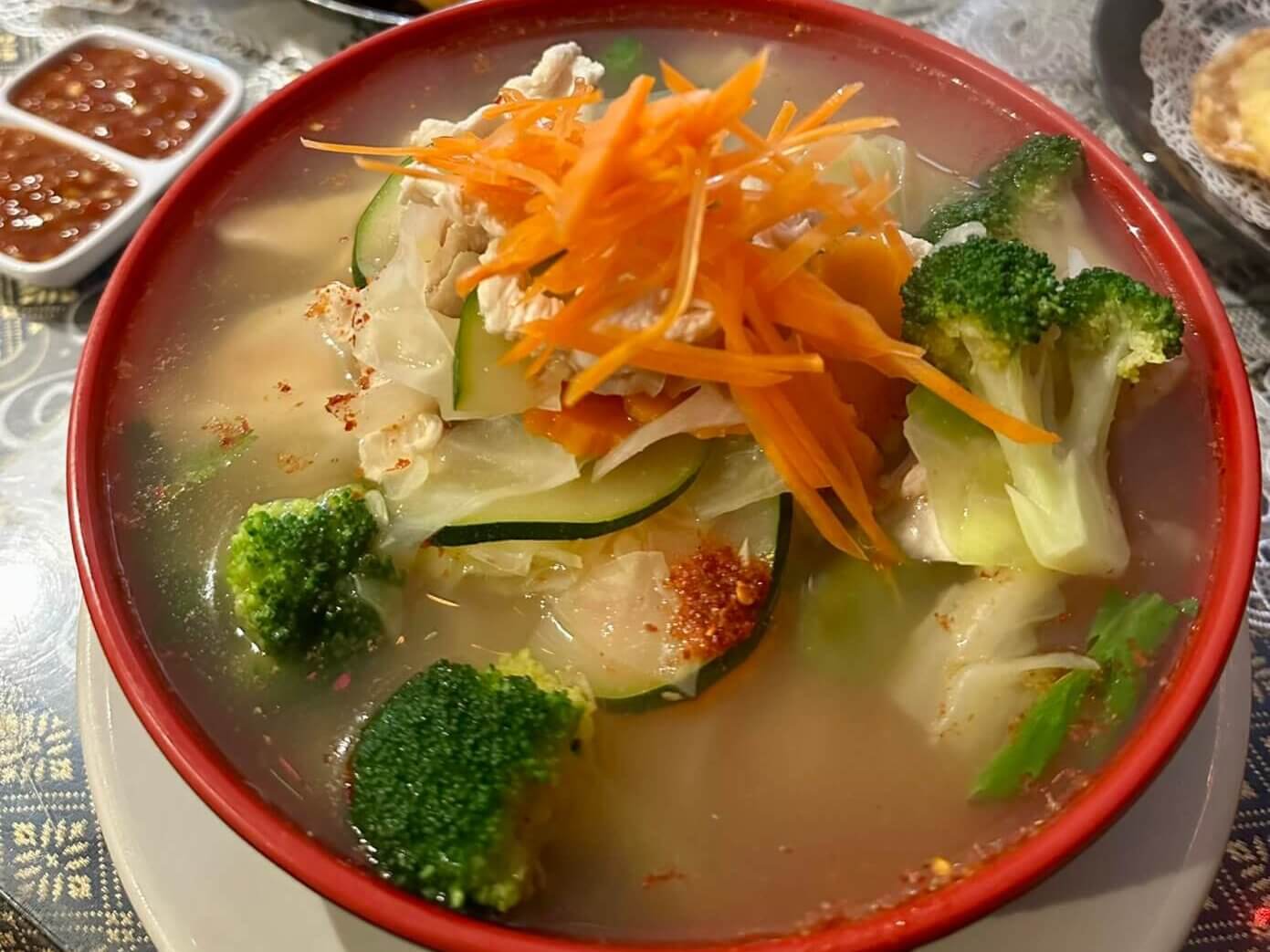 Red soup bowl with hot soup with a medley of vegetables, steamed chicken, and a garnish of sliced carrots.