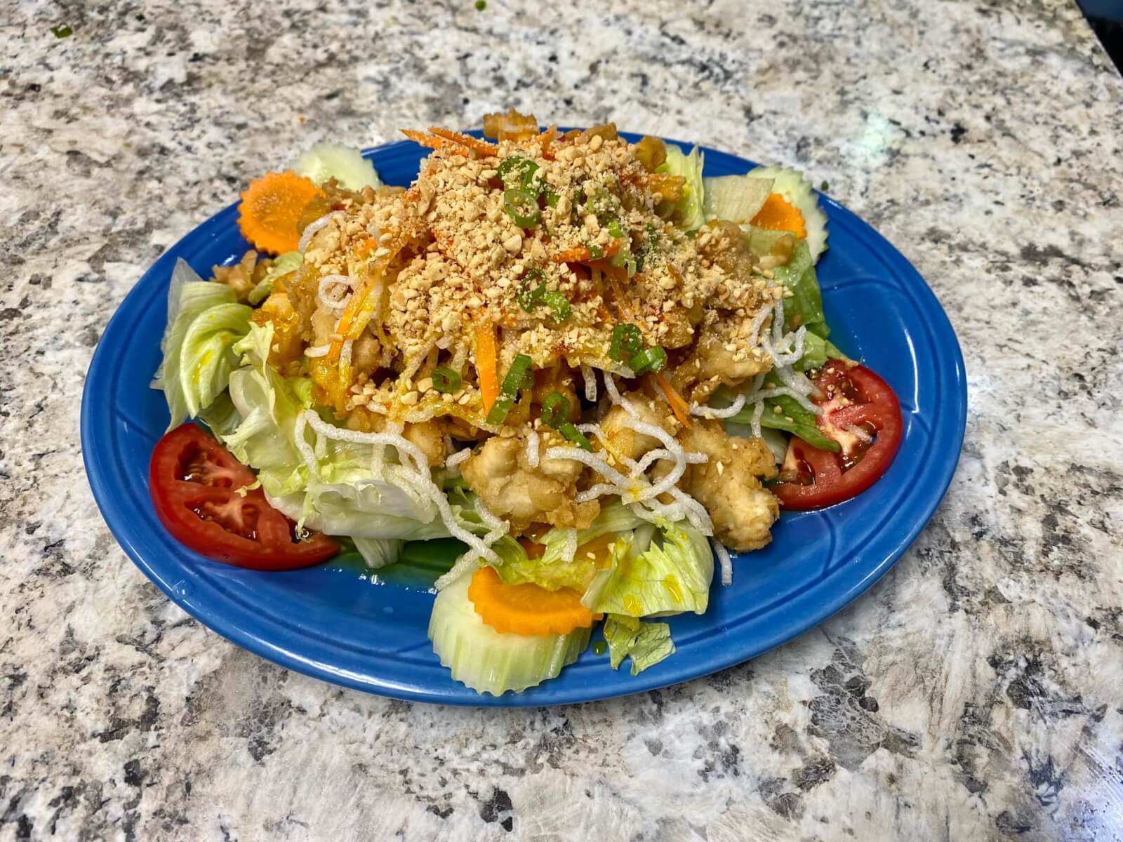 A blue plated with a serving of a salad dressed with carrots, cucumbers, tomatoes, fried chicken and a garnish of sliced carrots, scallions, and crushed peanuts.