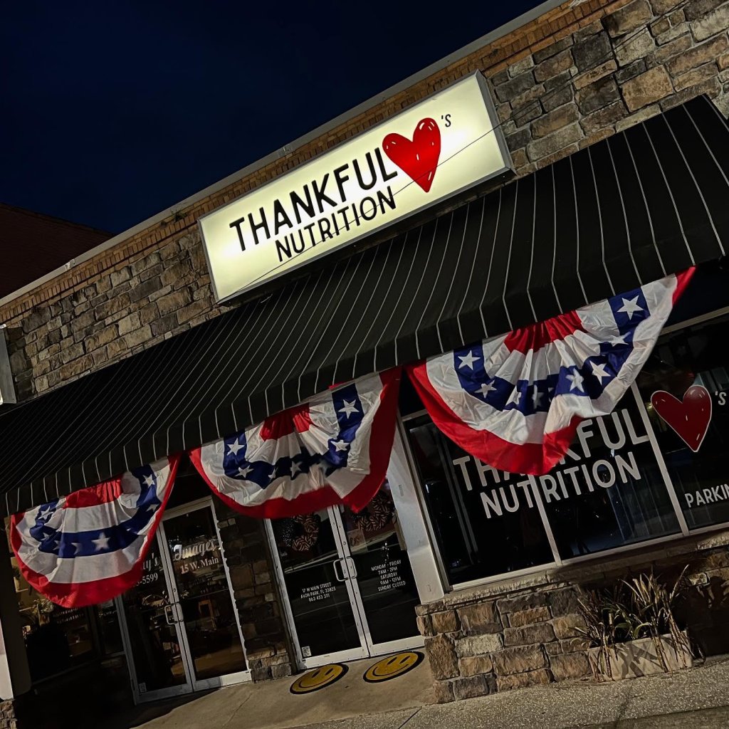 thankful-visit-sebring-fl A storefront in Avon Park, Florida, shown at night, with a sign reading “Thankful Nutrition” featuring a red heart. The building has a black striped awning decorated with red, white, and blue patriotic bunting, and the entrance has glass doors and stone exterior accents.