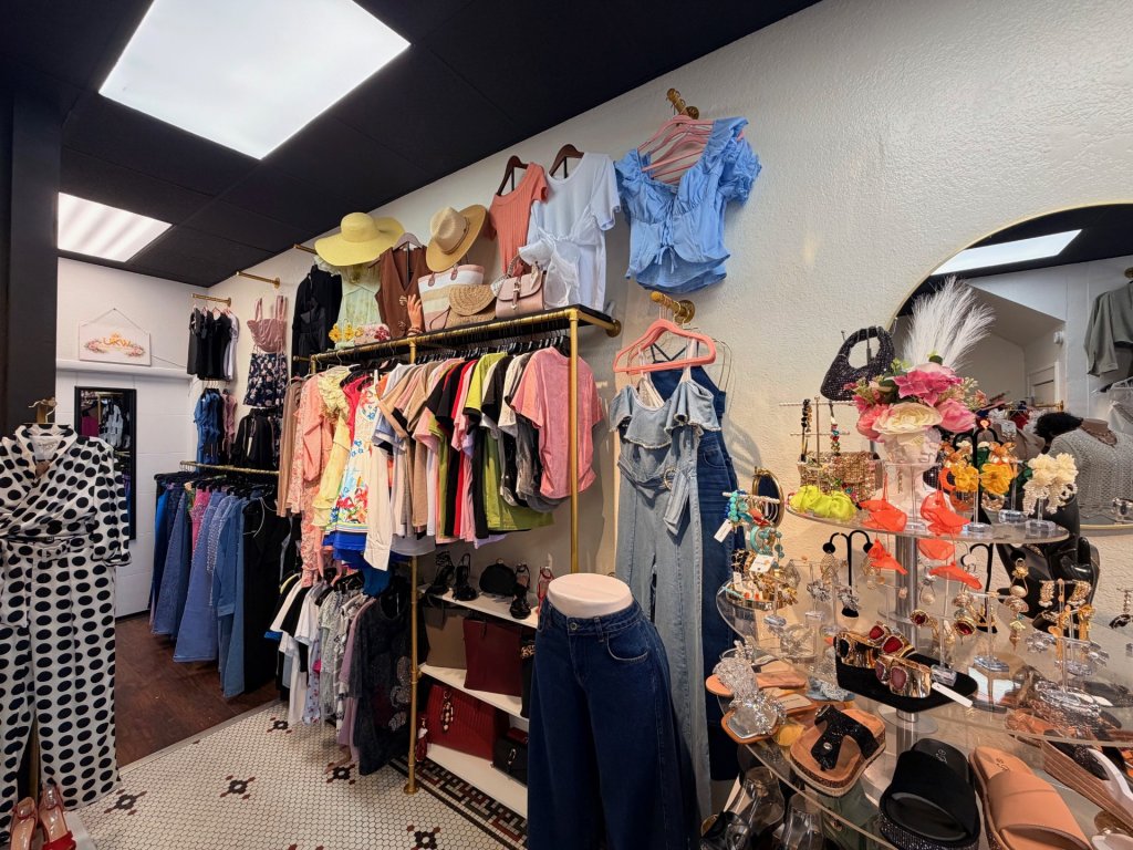 A clothes rack full of clothes, top racks with shirts, and in the front a display table with earrings, sunglasses, and shoes.