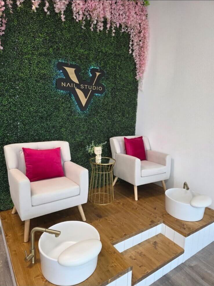 Green wall display with flowers hanging from the ceiling. The name of studio is shown against the greenery. There are two sofa chairs with hot pink pillows and footbaths directly in front of them.