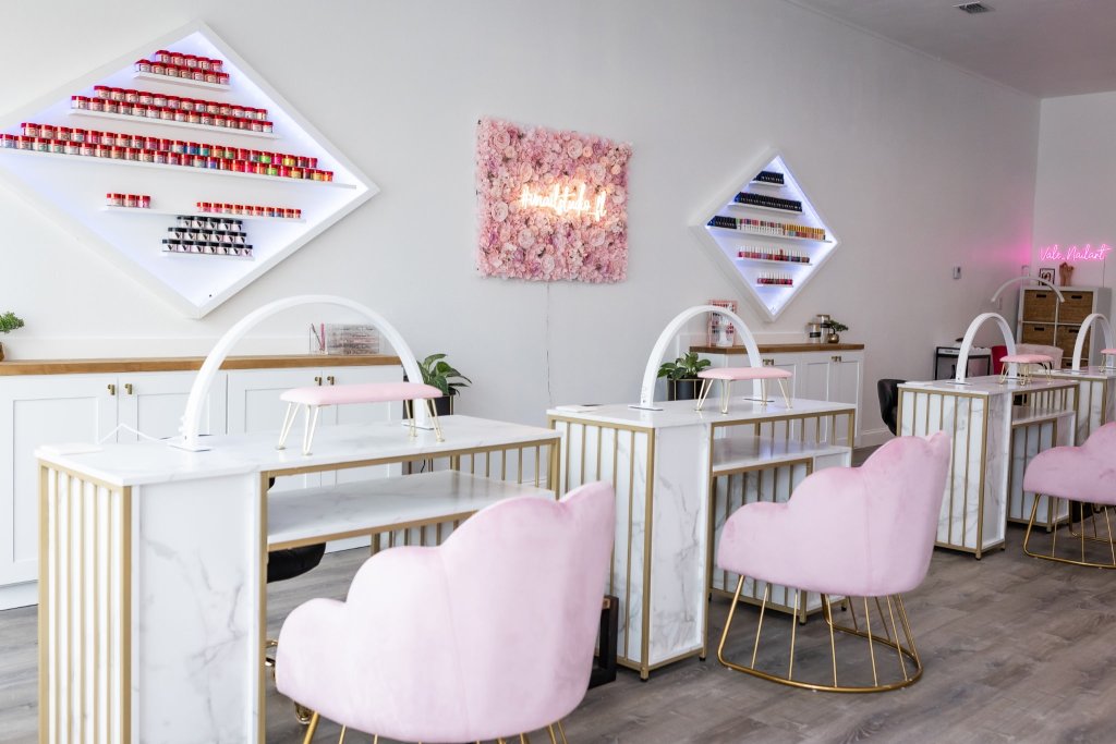 White painted nail studio in Sebring, Florida with pink accent chairs, floral portrait with neon sign and a display of all nail polishes.